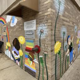 View from corner walls of the library. The mural features a rainbow with a young girl reaching up one side and another girl sitting on the ground reading. The other wall features kids and teens running and jumping with joy, two dogs, and backpacks bursting open with books coming to life. Dandelions growing and in bloom are throughout with seeds flying through the air carrying dreams.