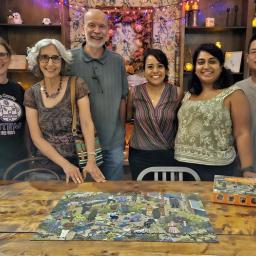 Six friends stand together smiling. In front of them is a table with a completed puzzle.
