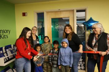 Adults and children in front of a closed door with red ribbon and scissors to conduct ribbon cutting and opening of the new sensory room.