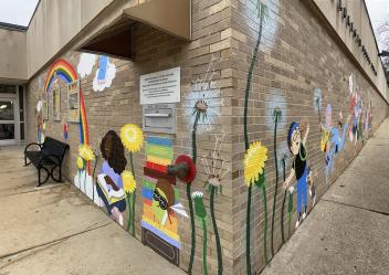 View from corner walls of the library. The mural features a rainbow with a young girl reaching up one side and another girl sitting on the ground reading. The other wall features kids and teens running and jumping with joy, two dogs, and backpacks bursting open with books coming to life. Dandelions growing and in bloom are throughout with seeds flying through the air carrying dreams.