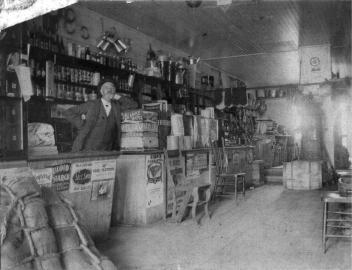 A black and white historical photograph of an early 20th-century general store. A man with a mustache, wearing a suit and hat, stands behind a wooden counter filled with various goods. Shelves behind him are stocked with bottles, tins, and other merchandise. The store is cluttered with wooden crates, barrels, and sacks. The ceiling is made of wood, and light streams in from a window or door on the right side.