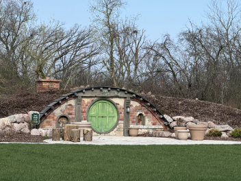 Hobbit-inspired berm built into a landscaped hill, featuring a round green door set in a brick-and-stone façade, with a curved roof, small windows, and a patio area with log seating in front, surrounded by grass and leafless trees.