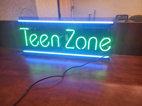 Picture of a lit neon sign that says Teen Zone in green with a blue line above and below the letters.