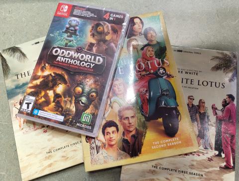 Photograph of three dvds, two copies of the first season of The White Lotus, one copy of the second season of The White Lotus, and one copy of a Nintendo Switch game Oddworld Anthology