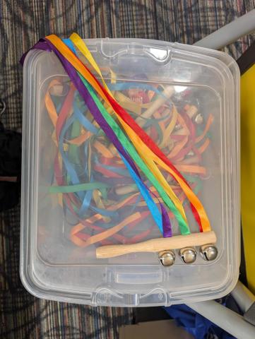 A single wooden handled bell instrument with three jingle bells and colored ribbons adhered to it.