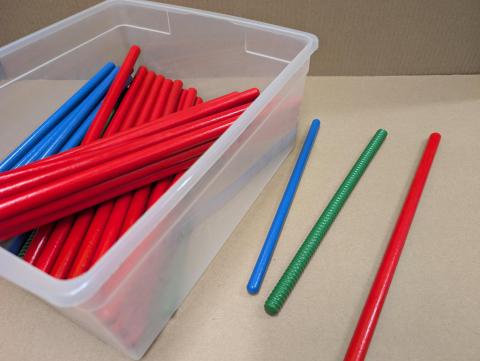 A plastic container, with a set of red, blue, and green rhythm sticks. One stick of each color rests beside the container.