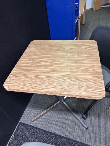Small square wood-grain table with rounded corners and a metal pedestal base. The table is in used condition with a small dent along one side.