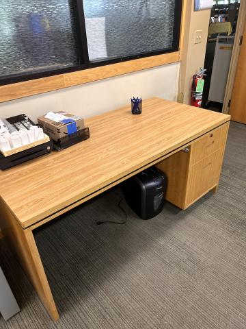 Large wooden office desk with a spacious rectangular work surface, built-in drawers on the right side, and sturdy wooden legs. The desk has a natural light oak finish and shows some signs of normal use.