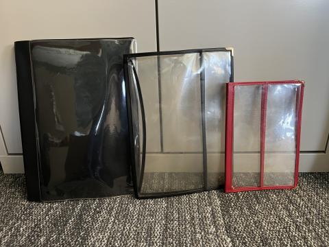 Three magazine covers in different sizes leaning against a cabinet on a carpeted floor. The covers are black, clear with black trim, and red with clear panels.