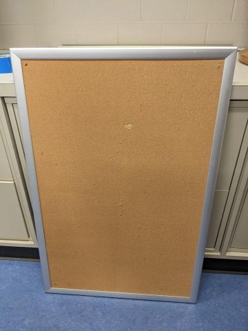 An aluminum lined cork board resting against a filing cabinet.