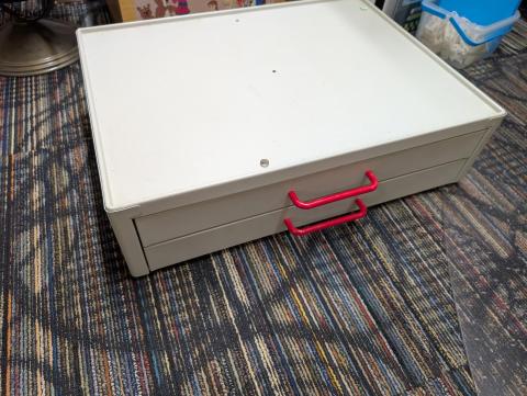 A beige set of drawers with red handles, resting on the carpeted ground.