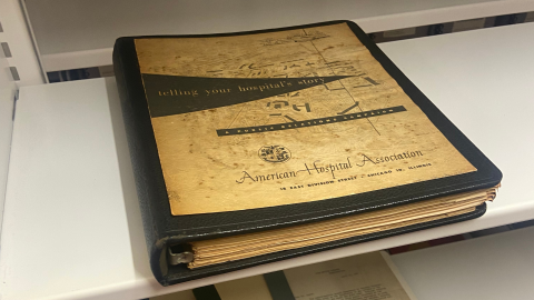 An aged, dark brown leather binder with a parchment cover sits on a metal shelf. The cover shows a map and the words, "Telling your hospital's story. A Public Relations Campaign. American Hospital Association, 18 East Division Street, Chicago 10, Illinois." 