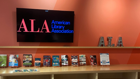 A television hanging on an orange wall displaying the letters A-L-A and the words American Library Association. Beneath the television are shelves displaying books on easels.