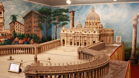 A miniature replica of St. Peter's Basilica on a display table. The walls behind the piece are painted with blue sky scenes from Rome.