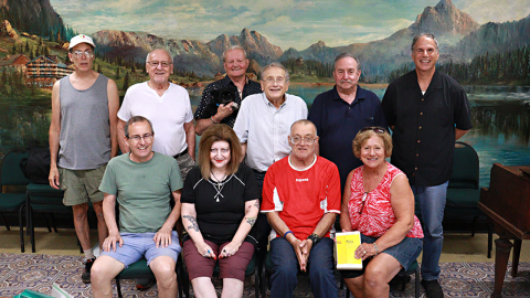 A group of 6 people stand behind 4 people sitting, all different ages. They smile into the camera with a scenic mural drawn on the wall behind them. 