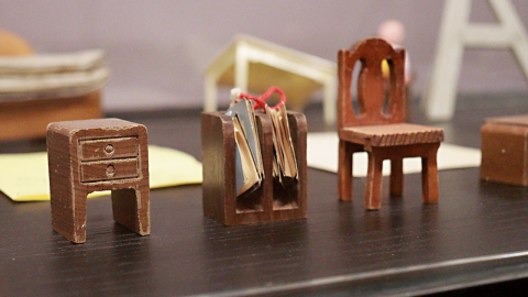 A miniature wooden table, bookshelf, and chair set in a glass display case. 