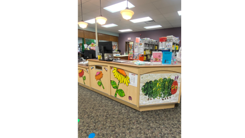 The circulation desk of an elementary school library. A picture of the Very Hungry Caterpillar hangs on the front, and bright lights hang down from overhead.