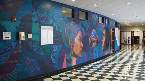 A mural painted on a wall depicting two people of color surrounded by abstract, colorful patterns. A black and white checkered floor lines the hallway.