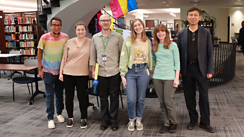 A group of six library workers stand in front of a spiral staircase in the Dominican University library, smiling into the camera. 