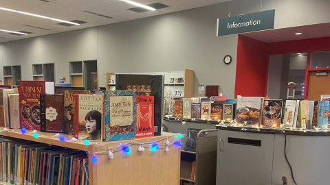 Rows of low, wooden bookshelves in a library. On top of the shelves, books are displayed standing up. A sign reading, "Information." hangs from the ceiling off to the right.