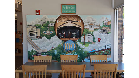 A centennial mural hangs on a wall behind a table with six chairs. The mural reads “Fox River Grove” and depicts many different town attractions, including a Metra train, a theatre, and ski jump.