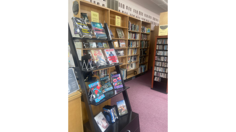 A row of wooden bookshelves full of library books line a wall. A single black bookshelf hoding books and movies in easels is in front of them.