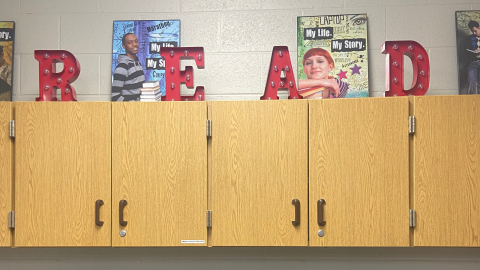 Wood cabinets hanging on a white cement block wall. On top of the cabinets are two posters showing people and the words, "My Life. My Story." In front of the posters are red marquee-style letters R, E, A, and D.