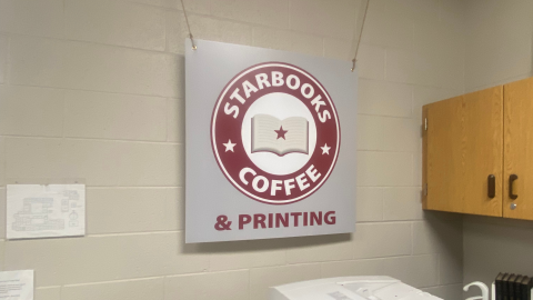 A grey sign suspended on ropes above a copy machine that reads, "Starbooks Coffee & Printing."