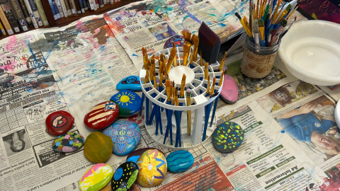 A table covered in old newspapers and in the middle of the table are a paintbrush carousel and a mason jar holding paintbrushes. On the left side of the table are painted rocks in various colors, and on the right is an empy white bowl. 