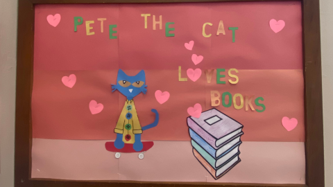 A bulletin board decorated with three shades of red paper arranged in stripes from dark to light. On the board are pink hearts, a stack of four books in red, green, blue, and purple, and a cutout of Pete the Cat. The text reads, "Pete the Cate loves books."