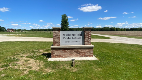 A brick and cement sign reading "Williamsfield Public Library, 407 Norman Drive" sits in a patch of green grass next to a concrete driveaway with a blue sky overhead.