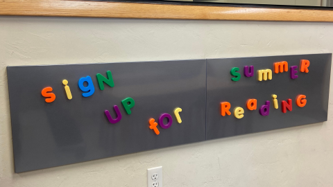 A dark grey magnetboard hanging on an off-white wall under an oak countertop. Magnetic letters on the board spell out, "Sign up for summer reading."