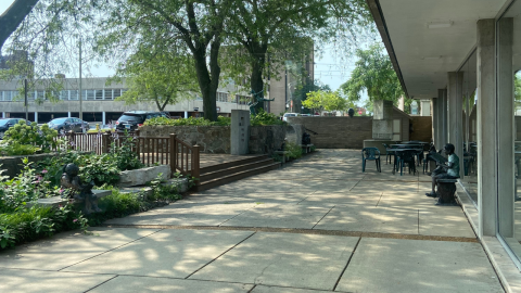 A courtyard area outside the library with foliage and steps on the left leading leading away from the library. Straight ahead, along the concrete terrace, a statue of a boy reading a book sits on bench. There are tables and chairs beyond the bench, and the window of the library are to the right. 