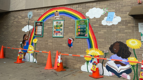 A mural of a child with dark brown skin holding a dandelion and standing to in front of two large dandelions. The child is to the left of a large rainbow with posters hanging in front of it. To the right of the rainbow, another child with dark brown skin is seating reading a book among dandlions.