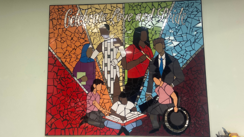 a ceramic tile mosaic with six sections in a rainbow color scheme from left to right. In the middle of the mosaic are 7 people representing various ethnic groups, ability groups, and genders. The top reads "Celebrate Love and Pride."