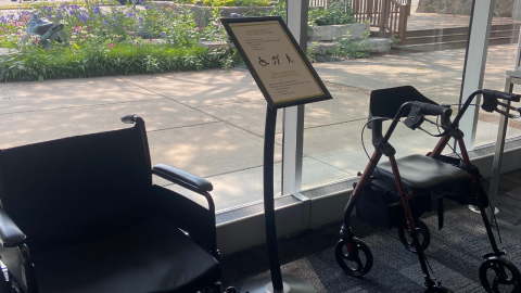 A sign promoting mobility accessibility at the library sits between a black wheelchair and a black rolling walker with a seat.