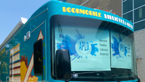 The front end of the Aurora Public Library Bookmobile, a bright aqua mobile book bus. The front of the bus above the windshield reads, "Boookmobile Biblioteca Movil." Two shades cover the windshield from the inside and read "Aurora Public Library District" with the library's logo.