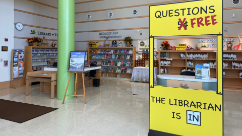 The atrium area inside the entrance to the library. There are shelves along the wall with wall signage indicating the Library of Things, Lucky Day Popular Materials, and the Patron Holds Pick-up area. In front of these shelves, are tables and an information desk. In front of the information desk is a large yellow cutout in the style of Charles Schulz's Peanuts comic stating "Questions FREE. The Librarian is IN." 