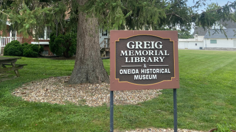 A dark brown sign with a light brown border mounted on two metal posts sits in front of a large tree with a white and red brick building in the background. The sign reads, " Greig Memoral Library & Oneida Historical Museum."