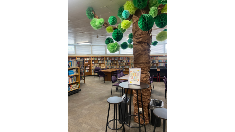 A post in the middle of the library wrapped in brown paper to resemble a tree trunk with green paper spheres handing from the branches to resemble leaves. The tree is surrounded by tables and chairs for library users, and there are bookshelves of books lining the walls beyond the tables. 