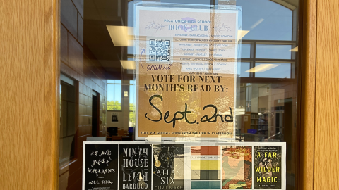 A yellow sign hanging in a window reads, "Pecatonica High School Book Club," and includes the dates and genres for upcoming meetings. On the left side of the sign is a QR code with the words "Scan Me" in red, and text at the bottom of the sign states the deadline to vote for next month's read, September 2nd.  Below the sign are images of the covers for various books. 