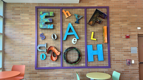 A metal letter collage hangs on a brown brick wall. The frame is purple and creates three sections featuring various metal letters in different colors and finishes.