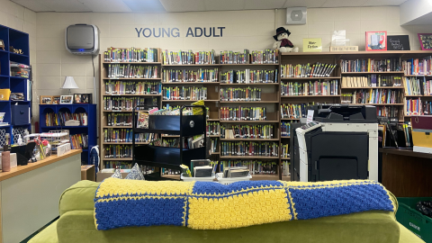 The young adult area of the library featuring shelves full of books along an off-white cement block wall. In front of the shelves are a black bookcart, a photocopier, the librarian's desk, and a greenish-yellow couch with a yellow and blue checkered afghan along the back.