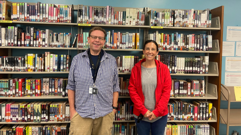 Two people stand in front of a tall bookshelf full of books; the person on the left is a smiling man with short brown hair wearing glasses, and on the right is a smiling woman with dark hair pulled back into a low ponytail. 