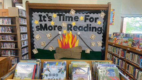 A bulletin board display behind rows of children's books. The bulletin board is covered in gray paper with a black border, and there are yellow, five-point stars and smores scattered across the display. It reads "It's Time for Smore Reading," in black letters above a construction paper campfire made of red, yellow, and organge flames, rising up from brown logs.