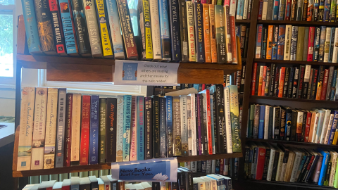 Brown wooden bookshelves full of various adult fiction books sits in front of a window. On the front are two signs, on the top shelf, a paper sign reads, "Check out what others are reading and then review for the next reader!" On the second shelf is another paper sign reading, "New Books. Just For You."