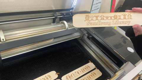 An open Glowforge laser cutter with several wooden bookmarks still lying on the tray inside the machine. At the right, a hand holds a bookmark decorated with a row of illustrated animal heads above the words, "Sandburg Library."