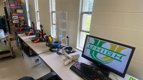 Two tables serving as the library's maker space hold four 3-D printers and spools of blue, yellow, and white plastic thread, as well as an AR/VR headset and computer. 