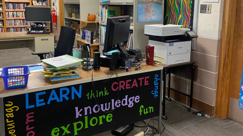 A tabletop circulation station holds a computer, stack of books, and a blue basket. To the right of the table is a photocopier on a stand, and behind the table is a tall, black rolling office chair. There is a second table behind the chair, with bookshelves along the far wall. 