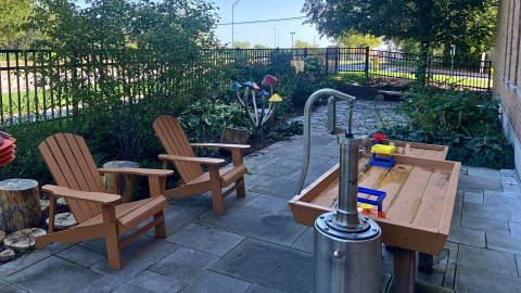 An outdoor waterplay area features a wooden water table with red, yellow, and blue children's toys. A silver metal water pump sits next to the table. To the left, are two brown, wooden adirondack chairs in front of green foliage and a black metal fence. The area is paved in dark grey slate pavers. 